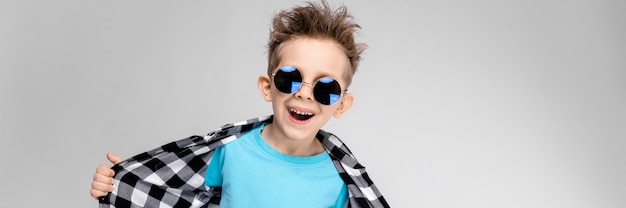 Photo a handsome boy in a plaid shirt, blue shirt and jeans stands. the boy is wearing round glasses. the boy smiles and pulls his shirt away
