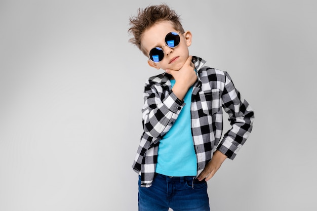 Photo a handsome boy in a plaid shirt, blue shirt and jeans stands. the boy is wearing round glasses. red-haired boy holds his hand to his chin