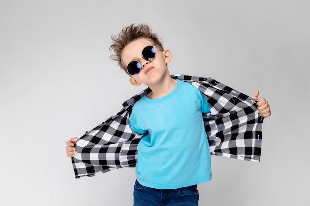 A handsome boy in a plaid shirt, blue shirt and jeans stands on a gray background. The boy is wearing round glasses. The boy smiles and pulls his shirt away