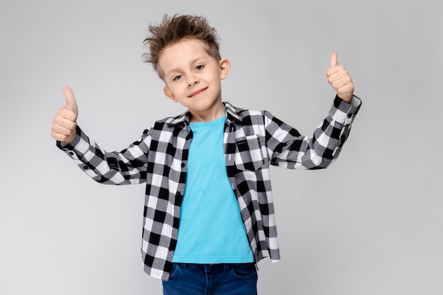 A handsome boy in a plaid shirt, blue shirt and jeans stands on a gray wall. 