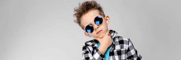 Un bel ragazzo in camicia a quadri, camicia blu e jeans si trova su una parete grigia.