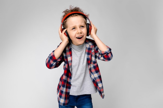 A handsome boy in a plaid shirt, gray shirt and jeans stands on a gray background. A boy in red headphones sings a song.