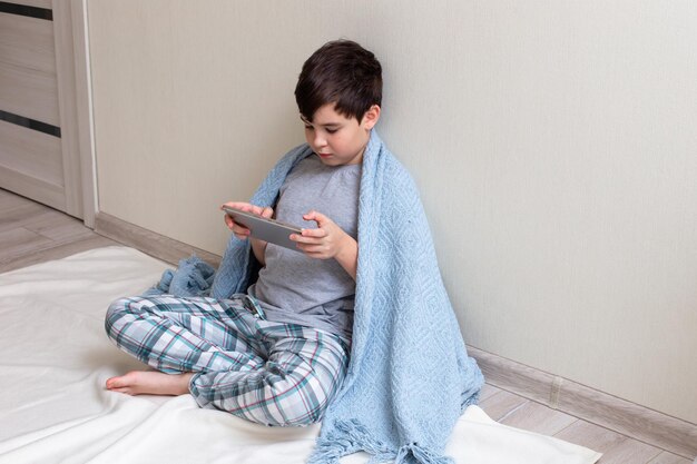 A handsome boy sits against a light wall on the floor holds a digital tablet