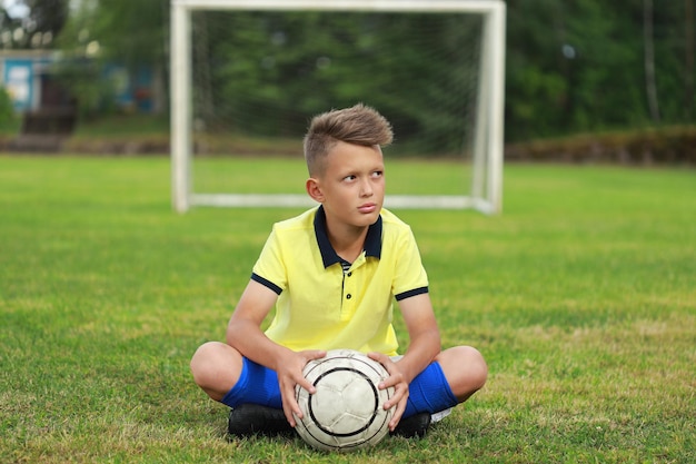 ハンサムな男の子のサッカー選手は、ボールでゴールを背景にサッカー場に座っています