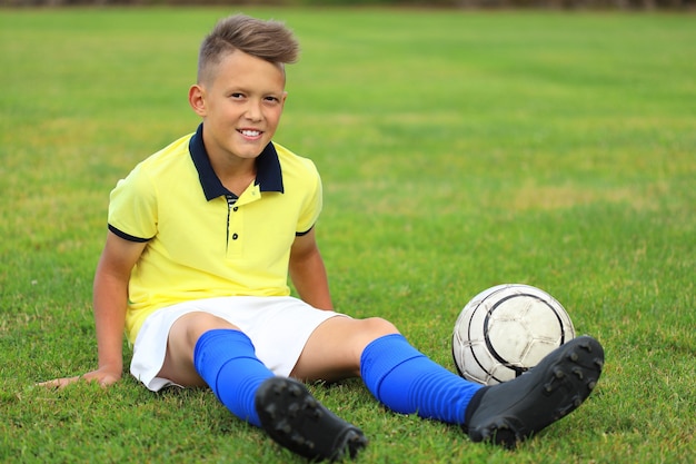 Красивый мальчик-футболист в желтой футболке и синей гантре сидит на футбольном поле