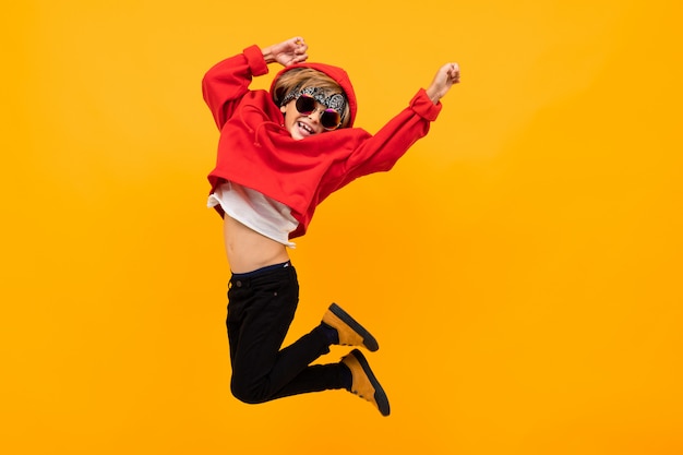 Photo handsome boy with a bandana on his head in a red hoodie with glasses jumps on isolated orange