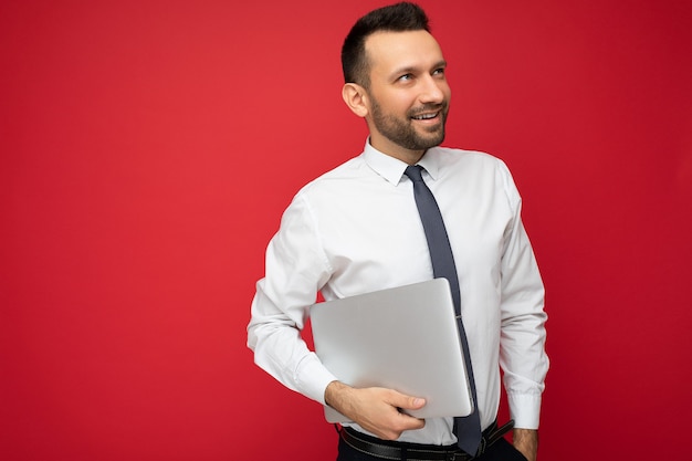 Handsome brunet man holding laptop computer looking to the side in white shirt and tie on isolated red background.