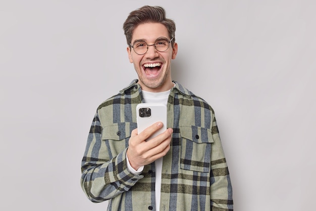 Handsome brunet man wears round spectacles and checkered shirt
uses smartphone sends sms scrolls internet laughs joyfully poses
against grey studio wall. people and modern technologies
concept