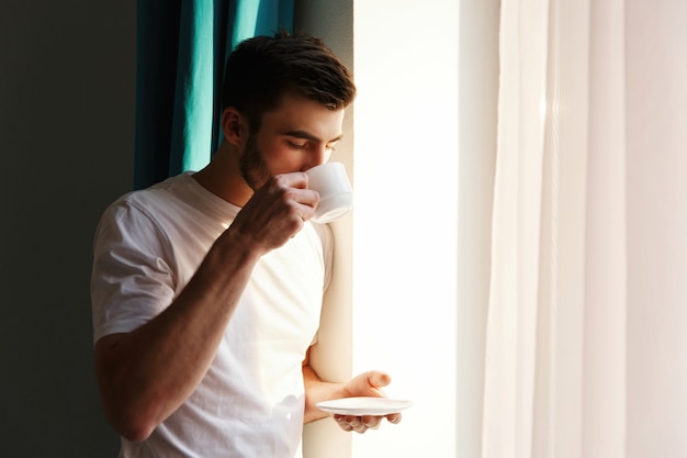 Handsome brunette bearded man stands before bright window and drinks coffee 