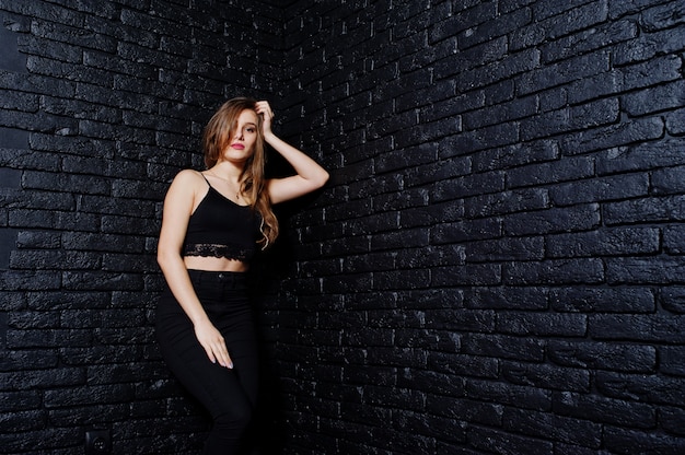 Handsome brunette girl wear on black, posing at studio against dark brick wall.