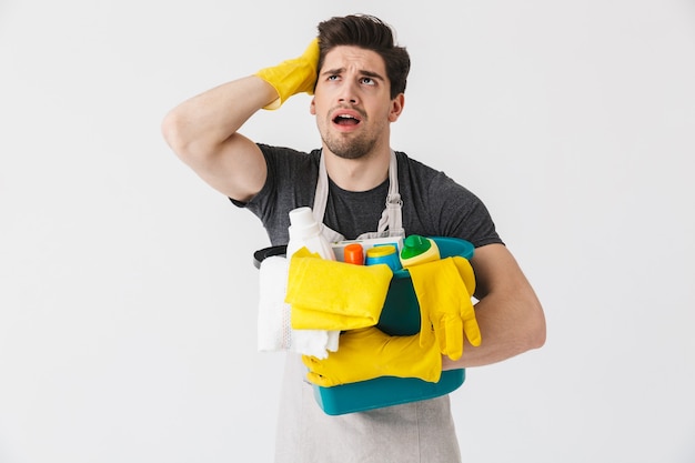 Handsome brunette houseman wearing apron standing isolated over white , holding bucket full of detergents