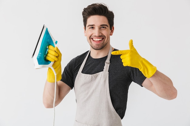 Handsome brunette houseman wearing apron standing isolated over white , showing iron