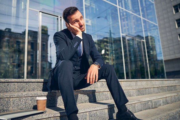 Handsome brunette male relaxing alone outside on stairs of business center with coffee