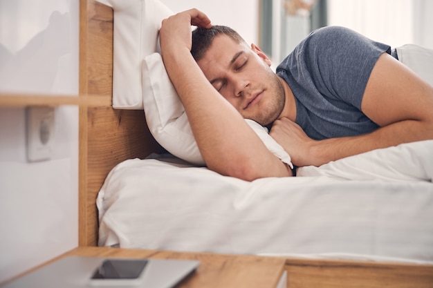 Handsome brunette male resting in comfortable bedroom on white blanket alone while seeing nice dreams