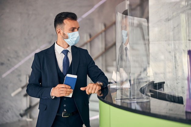 Handsome brunette man in a facial mask holding his passport while waiting to be checked at the custom