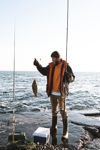 Bel pescatore barbuto brutale che indossa cappotto in piedi con una canna da pesca in riva al mare, mostrando pesce pescato