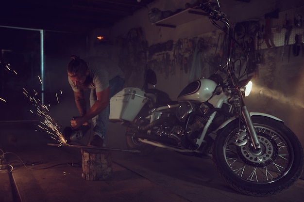 Handsome brutal male with a beard repairing a motorcycle in his garage working with a circular saw. In the garage a lot of sparks and smoke from sawing