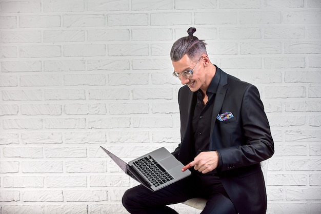 Handsome business man in glasses and suit holding laptop in hands and writing something. Side view. On white brick 