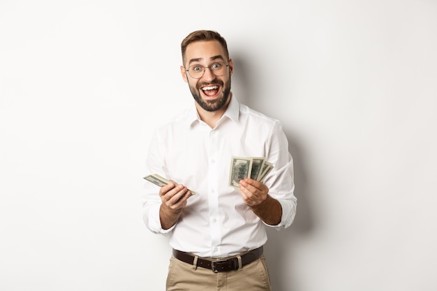 Handsome business man looking excited while counting money, standing over white background.