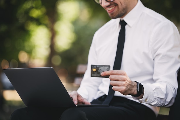 Handsome business man outdoors in the park using laptop computer holding credit card.