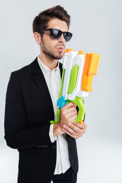 Handsome businessman in black suglasses holding colorful water gun over gray wall