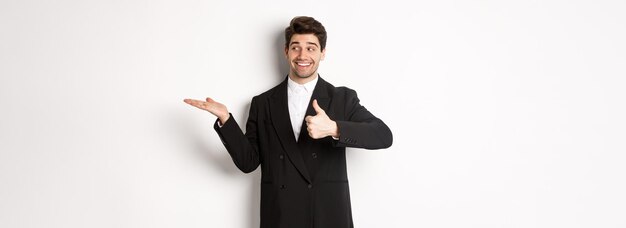 Handsome businessman in black suit showing thumbup and holding your product in hand over white copy