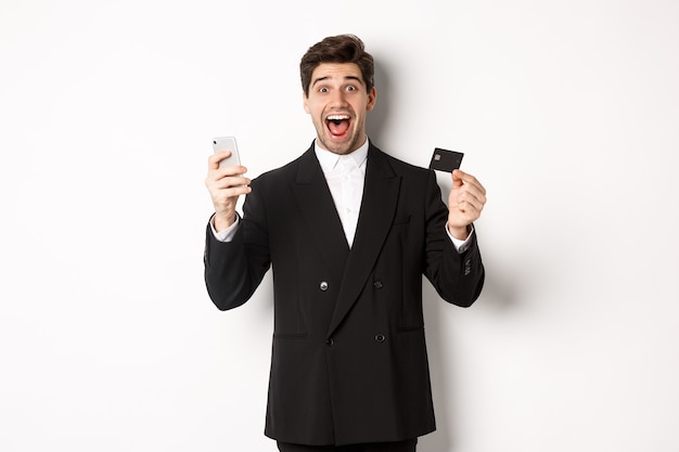 Handsome businessman in black suit smiling, showing credit card and money, looking amazed, standing against white background.