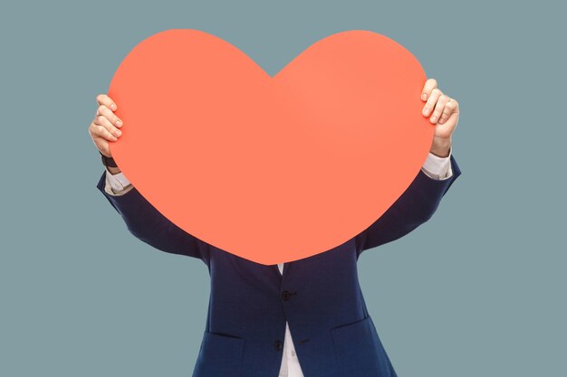 Photo handsome businessman in blue jacket standing and holding and covering red big heart shape in front of face for copy space. indoor, studio shot isolated on light blue background.