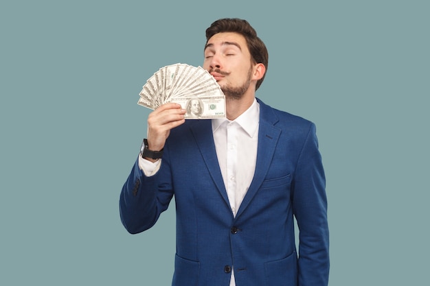 Handsome businessman in blue jacket standing and holding many dollars in hand and smelling and feeling smell of richness with closed eyes. Indoor, studio shot isolated on light blue background.