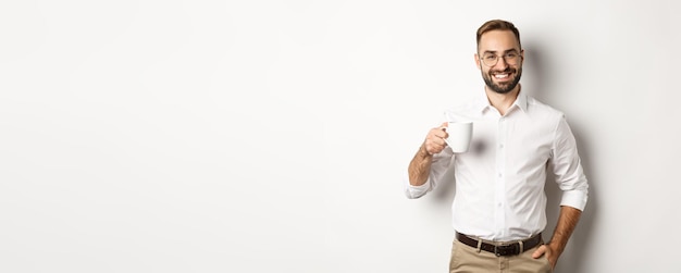 Uomo d'affari bello che beve caffè e sorridente in piedi su sfondo bianco