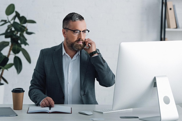 Handsome businessman in formal wear talking on smartphone and looking at computer