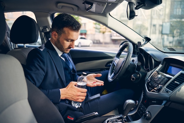 Handsome businessman holding pills in hand and water while spending time in auto