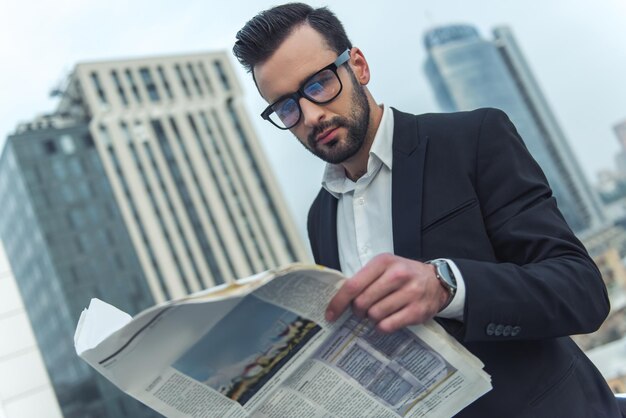 スーツと眼鏡のハンサムなビジネスマンは、バルコニーに立っている間新聞を読んでいます