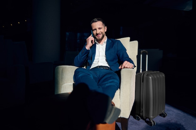 Handsome businessman, investor, sits in an armchair, resting in hotel lounge area with outstretched crossed legs on an ottoman, smiles talking on a cell phone, looking at camera during a business trip