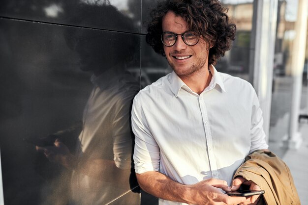 Handsome businessman leaning on a wall while standing outdoors and using smartphone on the street Happy man with curly hair wearing white shirt and beige coat in hand looking at his friends