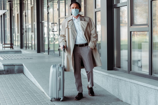 Handsome businessman in medical mask holding suitcase near building on urban street