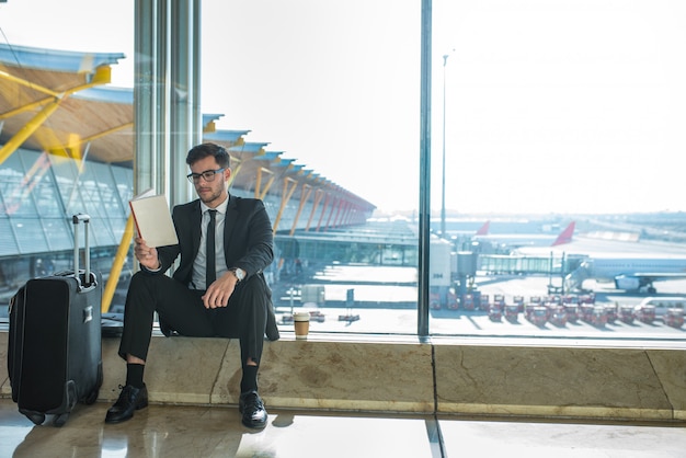 Handsome businessman reading a book in the airport sitting close to the window