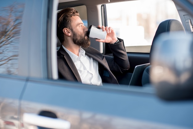 Handsome businessman sitting with coffee to go on the backseat of the car