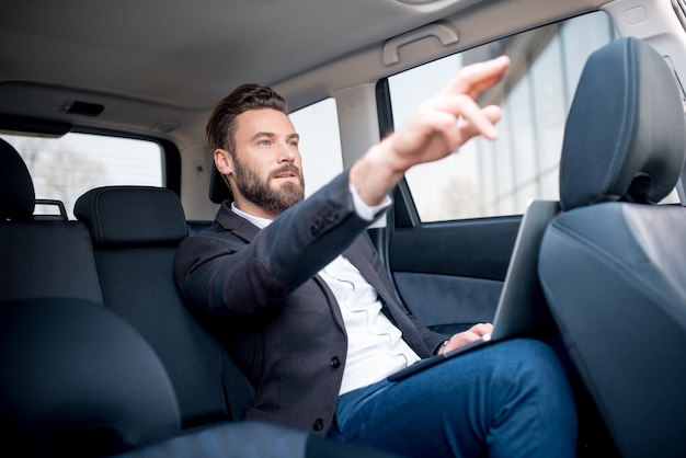 Handsome businessman sitting with laptop on the backseat of the car