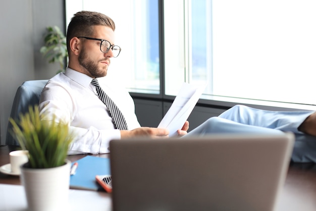 Handsome businessman sitting with legs on table and examing documents in office.