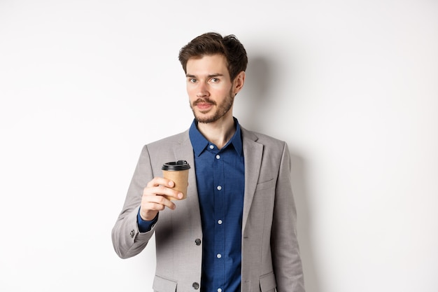 Handsome businessman in suit drinking coffee and looking at camera, standing against white background.