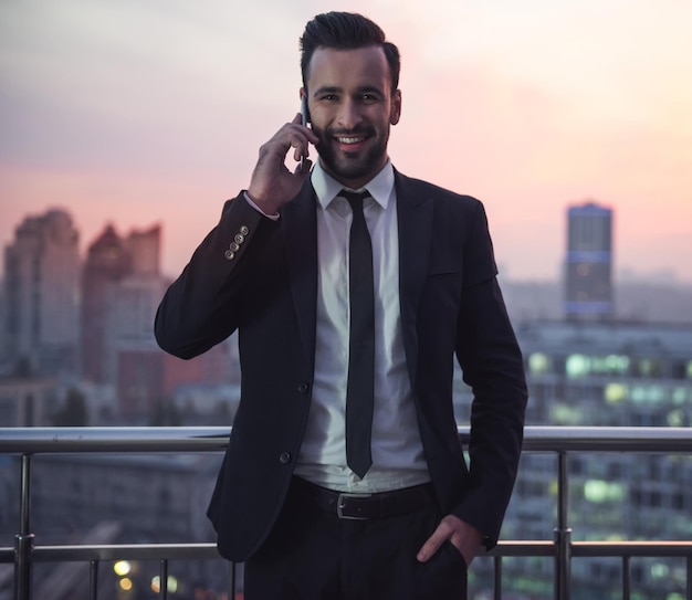 Handsome businessman in suit is talking on the mobile phone and smiling while standing on the balcony beautiful sunset behind him