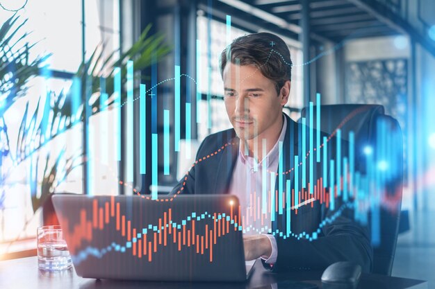 Handsome businessman in suit at workplace working with laptop to optimize trading strategy at corporate finance fund Forex chart hologram over office background