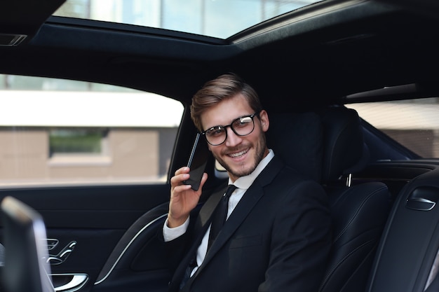 Handsome businessman talking with phone sitting with laptop on the backseat of the car.