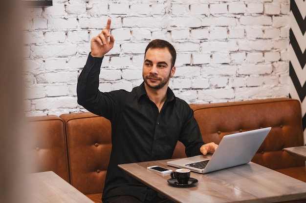 Uomo d'affari bello con il computer portatile che chiama cameriere mentre sedendosi in caffè urbano durante la pausa caffè