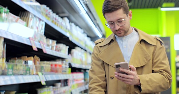 Handsome businessman with shopping trolley using smartphone food checklist walking inside grocery store