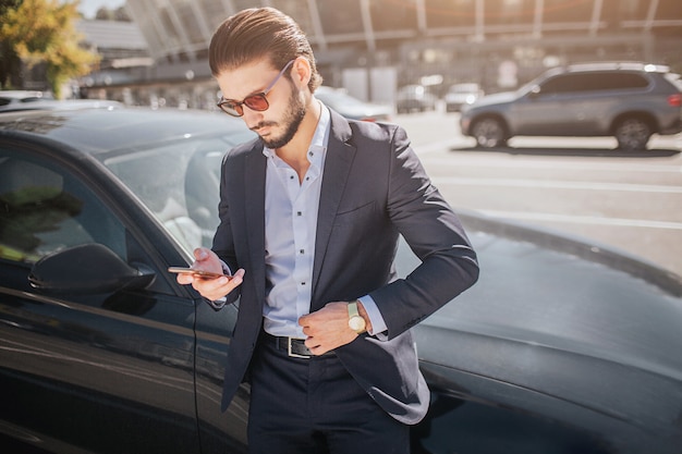 ハンサムで忙しい若い男は黒い車に立っています。彼は電話を持って見ています。外は晴れです。