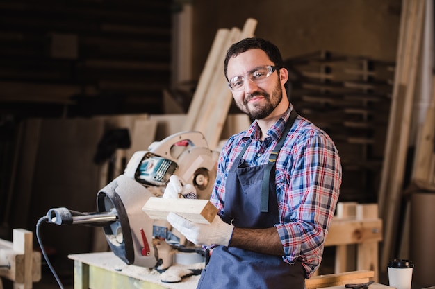 Handsome carpenter in protective glasses is looking at camera and smiling while standing near his wooden object in the workshop