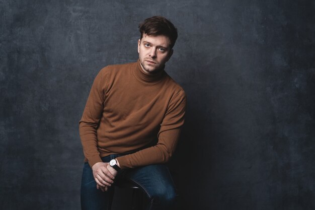 Handsome casual man sits on chair near a dark gray wall and looks to side.