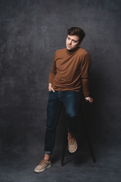 Handsome casual man sits on chair near a dark gray wall and looks to side.